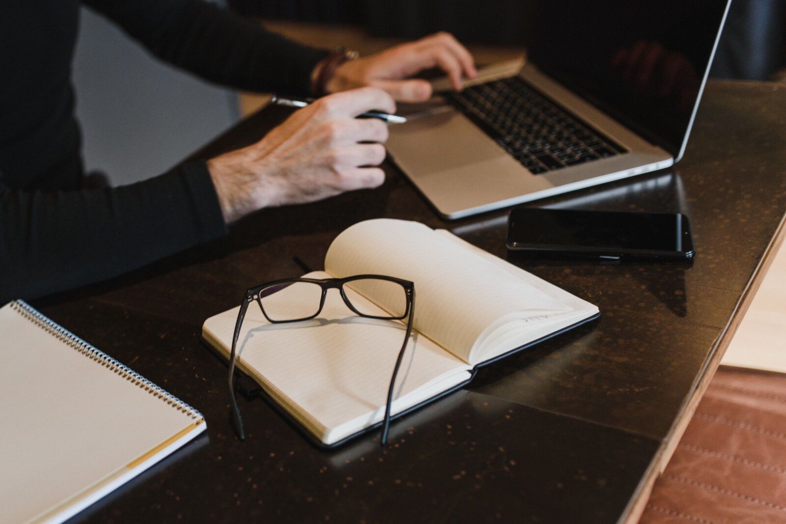 Hands using a laptop beside an open notebook and reading glasses.