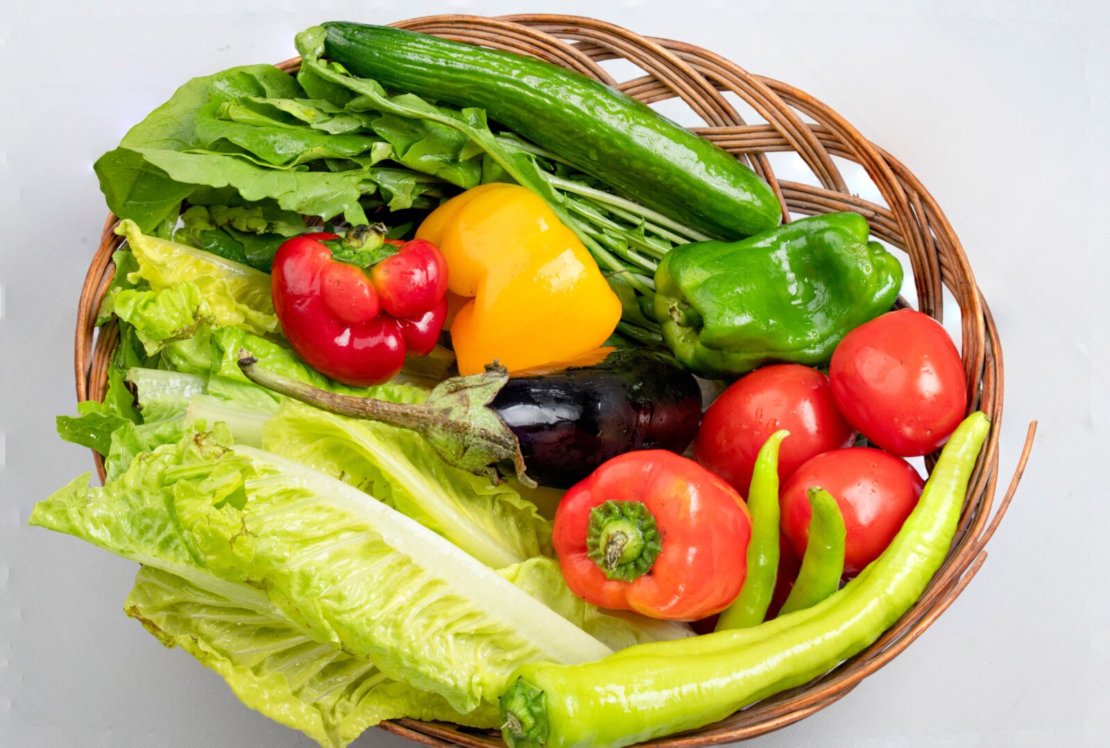 A wicker basket filled with peppers, tomatoes, cucumbers and leafy vegetables.