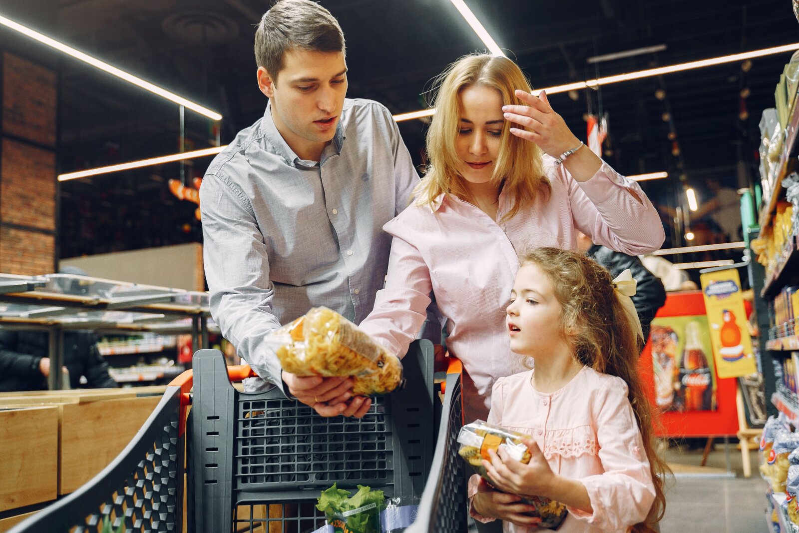 Two adults and a child selecting groceries beside a shopping trolley.