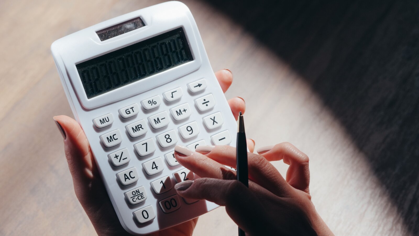 Hands holding a white calculator and pencil over a wooden desk.