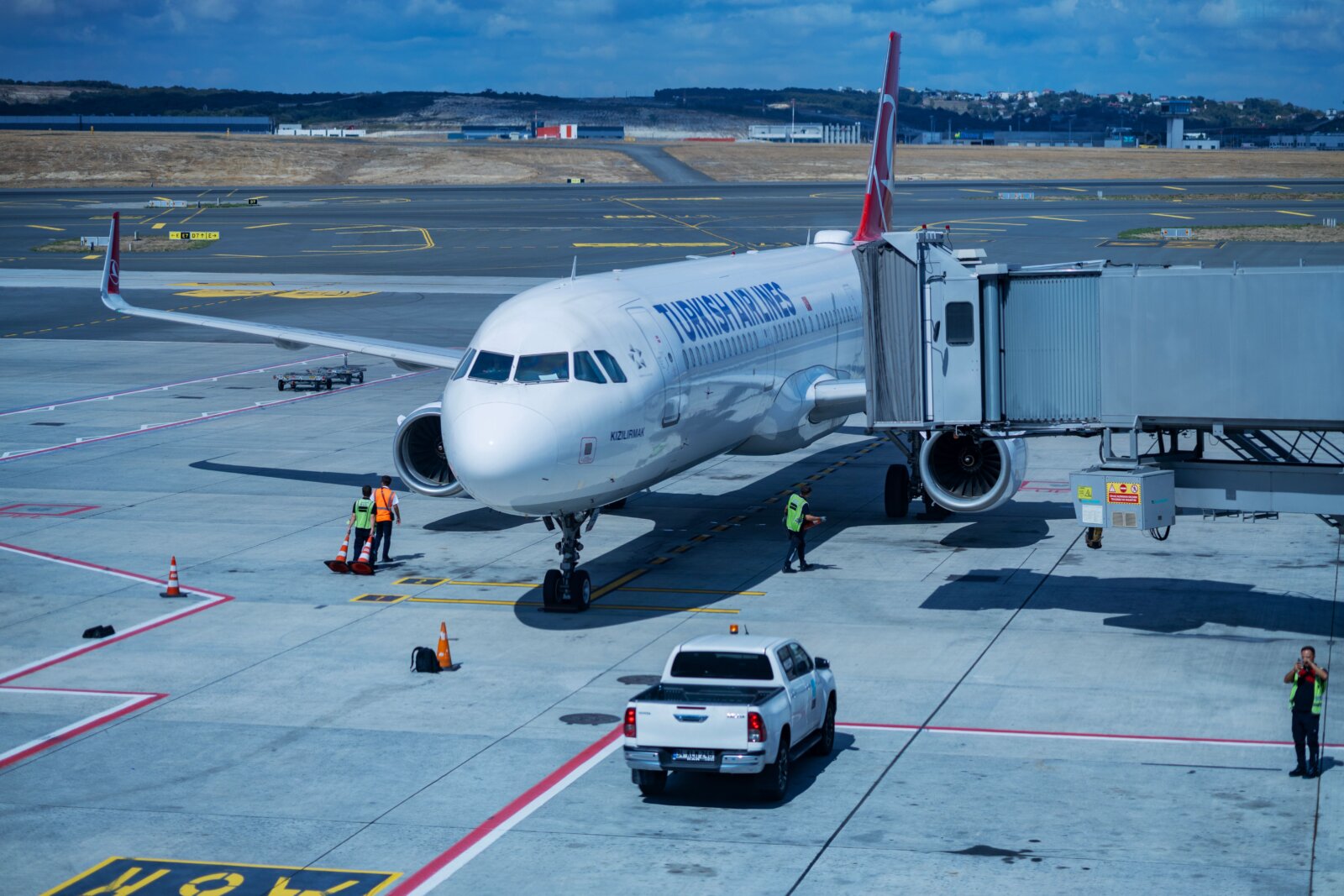A Turkish Airlines passenger aircraft beside an airport jet bridge with ground staff nearby.